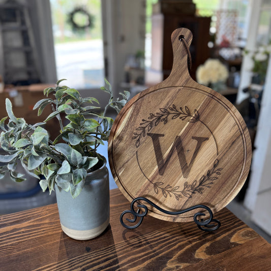 Wooden cutting board with engraved 'W' on a stand next to a potted plant on a wooden table.
